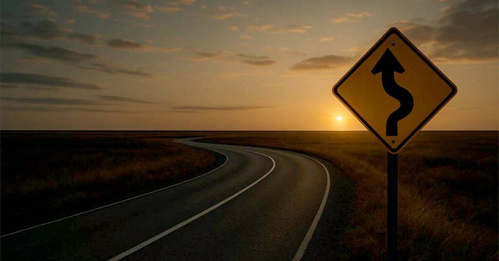 yellow warning traffic sign against winding road at sunset